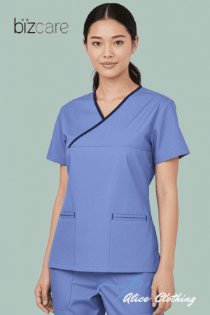 Young woman wearing blue medical scrubs with a wrap V-neck and two front pockets, standing against a pale green background.