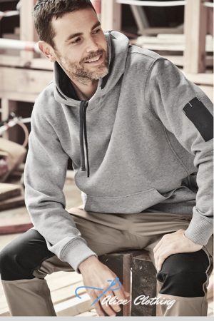Man with short dark hair and beard in a gray hoodie with a black sleeve patch, sitting on a wooden bench outdoors and smiling.