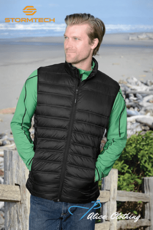Man modeling a black puffer vest over a green long-sleeve shirt on a beach, with Stormtech logo in the top-left and Alice Clothing watermark bottom-right.