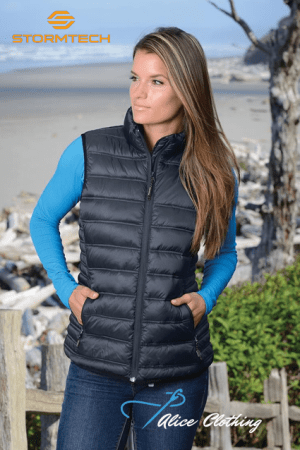 Woman wearing a black quilted vest on a beach with hands in pockets; Stormtech logo top-left and Alice Clothing watermark bottom-right.