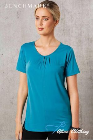 Model wearing a turquoise short-sleeve top with a subtle pleated neckline, standing against a gray textured background.