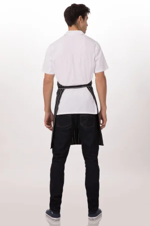 Back view of a man wearing a white short-sleeve shirt and a black striped apron tied at the waist, standing in jeans and sneakers.