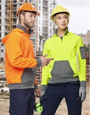 Two construction workers at a building site; man in orange hoodie and helmet gestures to the woman in neon-yellow jacket and yellow helmet.