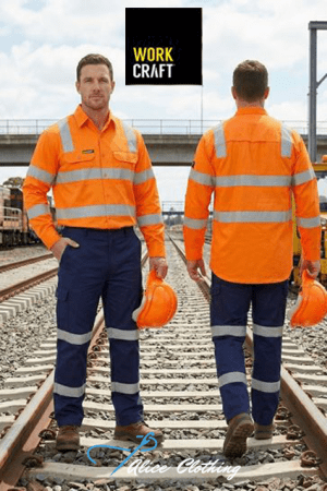Two construction workers in bright orange high‑visibility jackets with reflective bands stand on railroad tracks; one faces the camera, the other faces away, both holding orange helmets under a clear sky with a WORK CRAFT sign overhead.