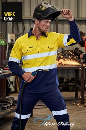 Welder in yellow-and-blue high-visibility coveralls lifts his helmet and holds a welding torch as sparks fly in a workshop.