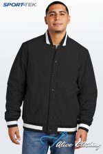 Man wearing a black varsity jacket with white striped cuffs and hem, standing against a light gradient background with SPORT-TEK logo in the corner.