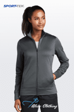Woman wearing a dark gray zip-up athletic jacket by Sport-Tek, standing against a light gradient background with the Sport-Tek logo visible in the corner.