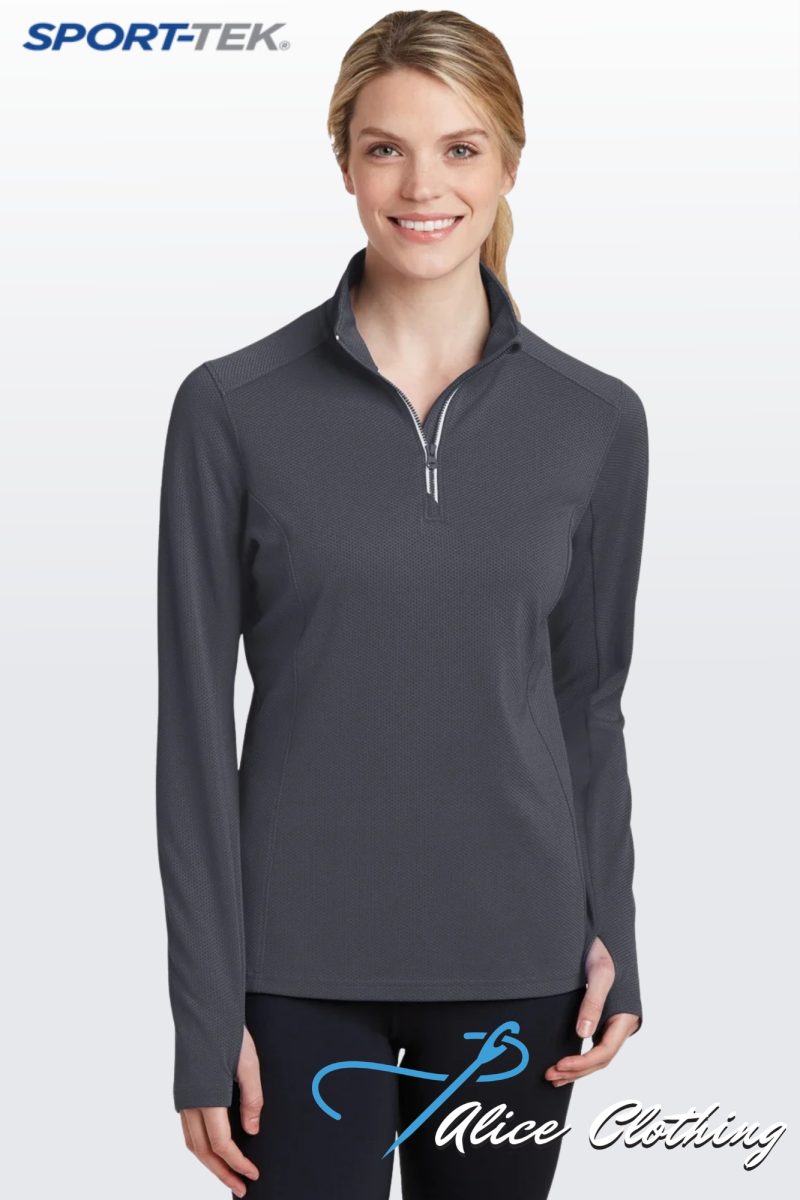 Smiling woman wearing a dark gray quarter-zip athletic top and black leggings with a blue script logo on the thigh; SPORT-TEK logo in the upper left corner.